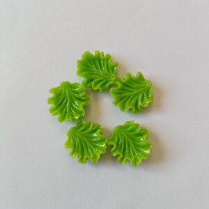 May include: Five bright green, leaf-shaped embellishments arranged in a circular pattern. Each leaf has a detailed, textured surface with prominent veins and a ruffled edge. The leaves are a vibrant, glossy green color.