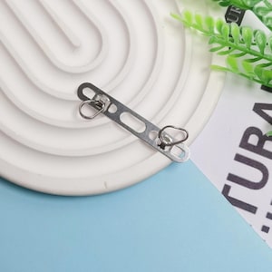 May include: A silver-colored metal clasp with two small loops and a series of holes. The clasp is positioned on a white, arched decorative piece, with a blue and white background.