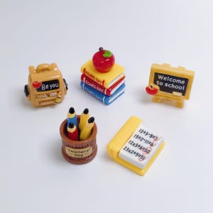 Op de afbeelding: Een set van vijf miniatuur schoolthema bedels van hars, inclusief een gele rugzak met de tekst 'Be you', een stapel van drie boeken met een rode appel, een geel schoolbord met 'Welcome to school', een bruine pennenhouder en een wiskundewerkblad.