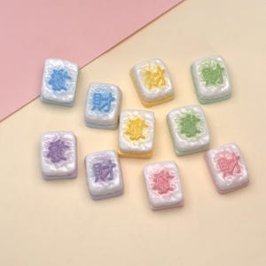 Puede incluir: Una colección de diez baldosas rectangulares en miniatura con diseños coloridos. Cada baldosa tiene una parte superior blanca con un símbolo de color, un borde de color y una base blanca. Las baldosas están dispuestas sobre un fondo rosa claro y amarillo.
