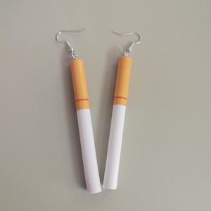 May include: A pair of novelty earrings designed to resemble cigarettes. Each earring features a white, cylindrical body with an orange-yellow tip and a red band. The earrings hang from silver-colored fish hook ear wires.