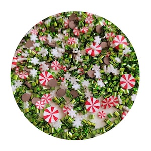 May include: A close-up of a white background with a variety of Christmas-themed sprinkles. The sprinkles include green and gold glitter, white snowflakes, brown chocolate discs, and red and white candy canes.