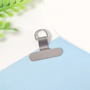 May include: A silver-colored metal clip with a rounded top and a flat, rectangular base. The clip is positioned on a light blue surface, with a white background and a touch of green foliage in the upper left corner.