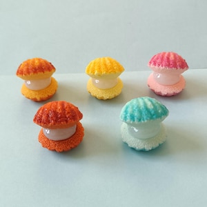 May include: Five colorful seashell-shaped decorative items. The shells are in shades of orange, yellow, pink, blue, and white. Each shell has a textured, ridged top and a smooth, round base. The items are arranged on a light blue surface.