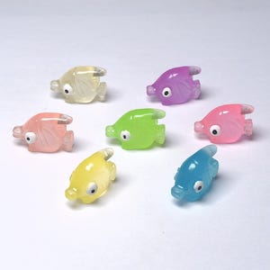 May include: A collection of translucent, colorful fish figurines in shades of yellow, pink, purple, green, and blue. Each fish has a white eye and a fin design. The figurines are arranged on a white surface.