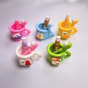 May include: Five miniature, colourful ceramic cups, each containing a toothbrush and toothpaste. The cups are pink, blue, yellow, white, and green, decorated with cartoon animal designs. The toothbrushes and toothpaste tubes are colour-coordinated.