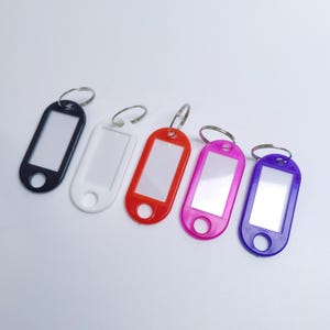 May include: Five oval-shaped plastic key tags with metal split rings, arranged in a row. Each tag features a rectangular white paper insert for labeling and a circular hole at the bottom. The tags are colored black, white, red, pink, and purple.
