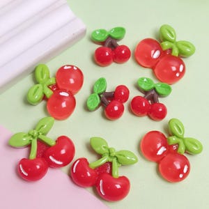 Puede incluir: Un conjunto de abalorios de cerezas rojas con hojas verdes y tallos marrones. Las cerezas son brillantes y parecen estar hechas de resina o un material similar. Los abalorios están dispersos sobre una superficie verde claro y rosa.