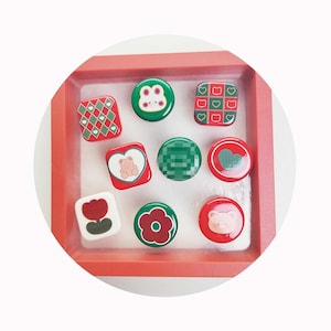 Könnte beinhalten: Neun kleine, runde, rote und grüne Knöpfe mit verschiedenen Designs, darunter ein Bär, eine Blume, ein Herz und ein kariertes Muster. Die Knöpfe sind in quadratischer Form auf einem weißen Hintergrund angeordnet.