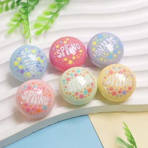 Op de afbeelding: Zes kleurrijke, ronde objecten in pasteltinten, waaronder roze, blauw, geel en groen, met het woord "SPRING". Het woord is omgeven door sterren. Ideaal voor lente-thema knutselen of decoratie.