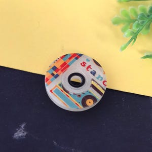 May include: A decorative tape dispenser with a colorful pattern and the word "stanc" printed on it. The dispenser has a clear center and a white outer ring. The design includes red, orange, blue, and yellow geometric shapes.