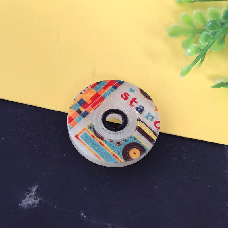 May include: A decorative tape dispenser with a colorful pattern and the word "stanc" printed on it. The dispenser has a clear center and a white outer ring. The design includes red, orange, blue, and yellow geometric shapes.
