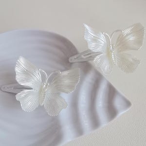 May include: Two white butterfly hair clips. Each clip features a detailed butterfly design with textured wings and a delicate body. The clips are made of a pearlescent material and are set on silver-toned metal clips. They rest on a white, scalloped surface.
