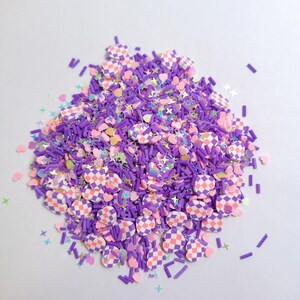 Puede incluir: Una mezcla colorida de adornos de arcilla polimérica. Incluye varillas moradas, corazones rosas, estrellas y círculos a cuadros rosas y blancos. El brillo iridiscente añade brillo.