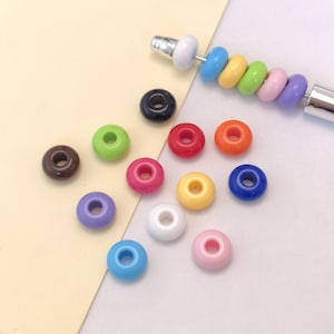 May include: A collection of colorful, round beads in various shades, including brown, green, pink, red, orange, yellow, blue, white, and purple. Some beads are strung on a silver-colored needle, while others are scattered around.
