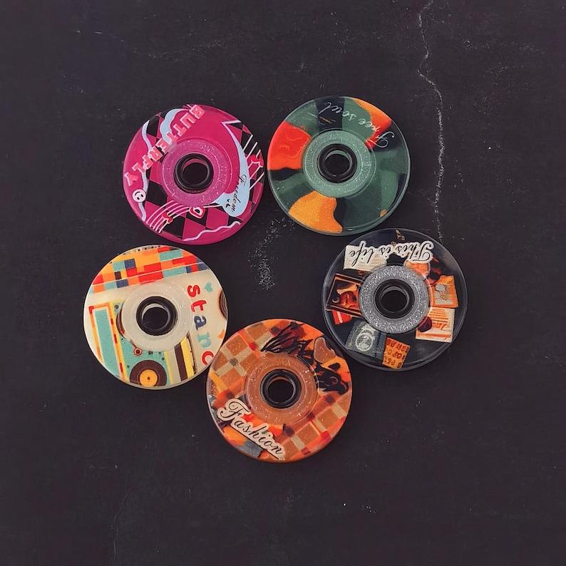May include: Five decorative bottle stoppers with unique designs. The stoppers feature various patterns, including floral, geometric, and text, in colors like pink, orange, green, and blue. Each stopper has a central hole.