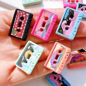 May include: A collection of colorful miniature cassette tape charms. Each charm features unique designs, including floral patterns, tropical scenes, and abstract art. The charms are in various colors, such as pink, blue, and black.