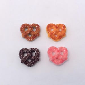 Può includere: Quattro ciondoli a forma di pretzel in miniatura disposti a quadrato. I pretzel sono in tonalità di marrone, arancione, marrone scuro e rosa. Ogni ciondolo ha una superficie strutturata, che suggerisce un alimento realistico.