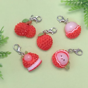 May include: A collection of red lychee fruit charms with silver-colored lobster clasps. Some charms are whole, while others are sliced, revealing the white fruit inside. Green leaves accent the charms, which are set against a light green background.