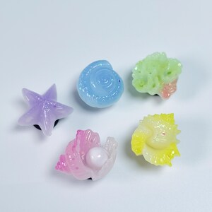 Peut inclure: Cinq petits ornements décoratifs aux formes marines : une étoile de mer lavande, un coquillage bleu clair, un morceau corallien vert pâle, un conque rose avec une perle blanche et un coquillage jaune en éventail, tous avec une finition brillante.