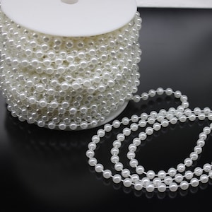 Puede incluir: Un carrete de guirnalda de cuentas de perlas blancas. La guirnalda está hecha de pequeñas cuentas redondas que están enhebradas en un hilo transparente. La guirnalda está enrollada alrededor de un carrete blanco.