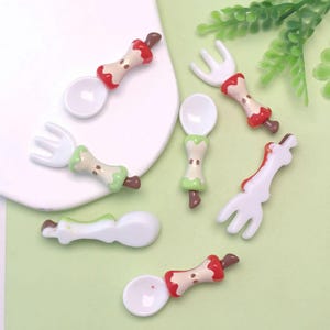Puede incluir: Una colección de cucharas y tenedores en miniatura con un diseño caprichoso de corazón de manzana. Las cucharas y tenedores tienen cuencos y púas blancos, con mangos de corazón de manzana en rojo, verde y beige. Los utensilios en miniatura están dispuestos sobre una superficie blanca y verde.