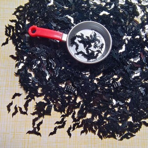 Op de afbeelding: Zwarte vleermuisvormige pailletten verspreid over een lichtgele ondergrond. Een kleine, zilveren maatbeker met een rode handgreep bevat meer pailletten. De pailletten zijn reflecterend en lijken van plastic te zijn.