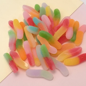 Puede incluir: Un montón de gusanos de goma coloridos en varios tonos de rosa, verde, naranja, amarillo y azul. Los caramelos tienen una capa de azúcar y están dispuestos sobre una superficie rosa claro.