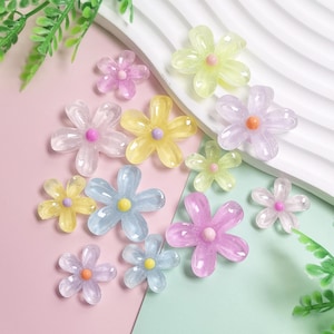Puede incluir: Una colección de adornos translúcidos en forma de flor colorida. Las flores son en tonos de rosa, amarillo, azul y transparente, cada una con un centro contrastante. Están dispersas sobre una superficie rosa y blanca.