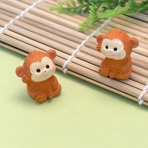 Puede incluir: Dos pequeñas figuras de mono naranja con caras de color beige claro y ojos oscuros. Los monos están sentados sobre una estera de bambú con follaje verde en el fondo. Las figuras son detalladas y parecen estar hechas de resina o un material similar.