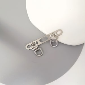 May include: A silver-colored metal bag hardware piece with two D-rings and a rectangular bar with a slot and two holes. The hardware is designed for attaching straps or other accessories to bags or purses. The piece is shown against a white and gray background.