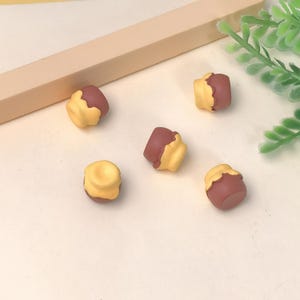 Pode incluir: Cinco mini pingentes de cupcake em amarelo e marrom escuro. Os pingentes estão dispostos sobre uma superfície bege clara, com um detalhe retangular marrom claro e um raminho de planta verde ao fundo.