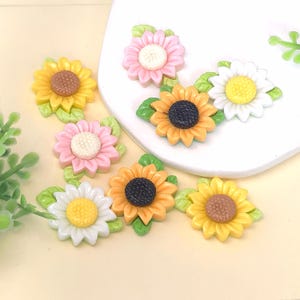 Può includere: Una collezione di abbellimenti colorati in resina a forma di girasoli e margherite. I fiori presentano petali gialli, arancioni, rosa e bianchi con centri a contrasto. Ogni fiore ha foglie verdi.