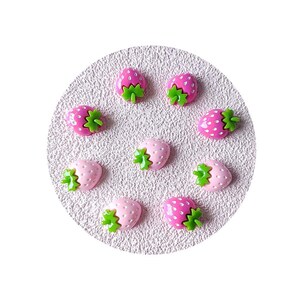 May include: A collection of nine strawberry-shaped embellishments. The strawberries are pink and fuchsia with green stems. They are arranged on a light pink, textured, circular surface.