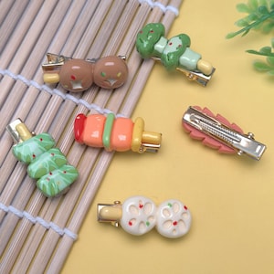 May include: A collection of colorful hair clips with various food-themed designs. The clips feature miniature representations of items like a tree, a leaf, and a skewer, all attached to metal barrettes. The color palette includes green, pink, orange, and brown.
