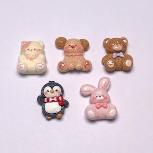 May include: A collection of five miniature resin figurines. The figurines include a white and pink cat, a tan dog, a brown bear, a black and white penguin with a red scarf, and a pink bunny with a bow.