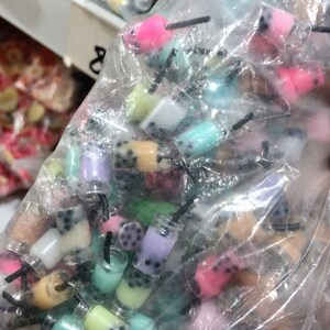 May include: A plastic bag filled with colorful miniature bubble tea charms. The charms are made of plastic and have a variety of colors, including pink, blue, green, and yellow. The charms are shaped like bubble tea cups with black straws.