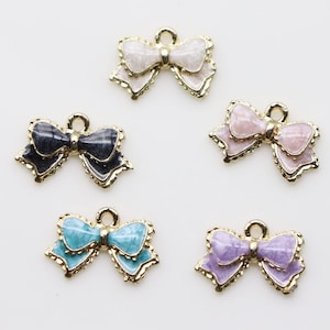 Op de afbeelding: Vijf gouden strikcharms met verschillende geëmailleerde afwerkingen. De strikken zijn in de kleuren wit, zwart, roze, turquoise en paars.