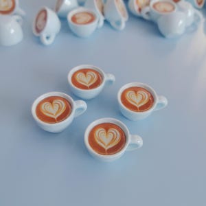 May include: Four miniature light blue ceramic coffee cups with brown coffee and a white heart design on the top of the coffee.