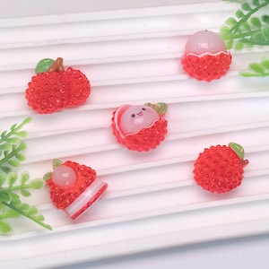 May include: A collection of miniature, detailed lychee fruit charms. The charms are primarily red with textured surfaces, featuring green leaves and white and pink inner details. Some are whole, while others are cut open, revealing the fruit inside.