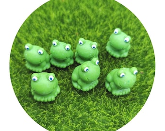 Puede incluir: Siete pequeñas figuritas de rana verde con ojos blancos, sentadas en un lecho de hierba artificial verde.