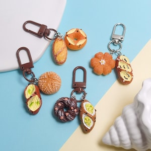 May include: A selection of keyrings with miniature food designs. The keyrings depict various baked goods, including bread, bagels, and a flower-shaped pastry. Each keyring has a brown clasp and ring.