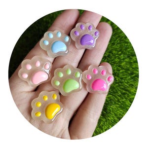 May include: Six colourful paw-shaped resin embellishments. Each paw features a different colour: blue, purple, pink, green, yellow, and pink. The paws are translucent with a glossy finish, ideal for craft projects.