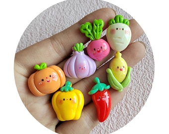 May include: A set of nine colorful, hand-painted, resin food charms. The charms are shaped like various vegetables and fruits, including a purple onion, a pink radish, a yellow pepper, a green corn cob, a red chili pepper, a white turnip, a yellow pumpkin, and a green garlic. Each charm has a cute, cartoon-like face.
