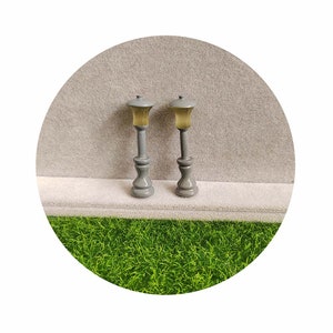 Peut inclure: Deux lampadaires miniatures gris avec des abat-jours en verre jaune. Les lampadaires sont sur une surface blanche avec de l'herbe verte en arrière-plan.