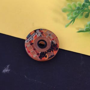 May include: A round, decorative button with a central hole. The button features a brown and orange checkered pattern with a horse silhouette and the word "Fashion" in white script. The button is set against a yellow and black background.