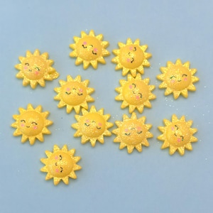 Könnte beinhalten: Eine Ansammlung gelber, sonnenförmiger Verzierungen mit lächelnden Gesichtern und Glitzerakzenten. Die Sonnen haben ein Sternmuster und sind auf einer hellblauen Oberfläche angeordnet. Diese dekorativen Elemente eignen sich für Bastelarbeiten.