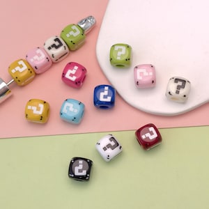 May include: A selection of colourful, cube-shaped beads, each displaying a white question mark on a coloured background. The beads are in various colours, including yellow, pink, blue, green, and black. Some beads are strung together, others scattered.