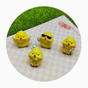 Puede incluir: Cuatro figuritas de pollitos amarillos con diferentes accesorios. Un pollito lleva auriculares, otro lleva gafas de sol, otro lleva una bebida y otro lleva un micrófono.