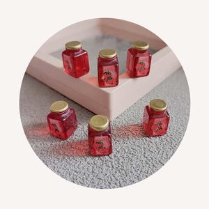 May include: Six small, square glass jars filled with red jam, each with a gold lid. The jars are labeled with the word "JAM" and a strawberry illustration. The jars are arranged on a light pink tray.